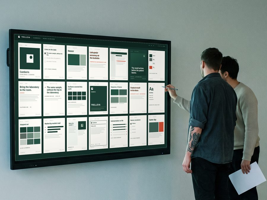 Two people at a lit wall display in a quiet room, reading finished work for one brand. One is marking a panel with a stylus.